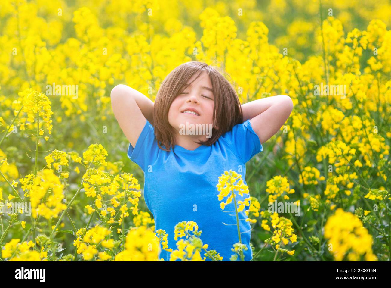 Bambino di cinque anni in una posa rilassata con le braccia sulla testa in mezzo a un prato di fiori di colza gialli durante una splendida giornata primaverile. Foto Stock