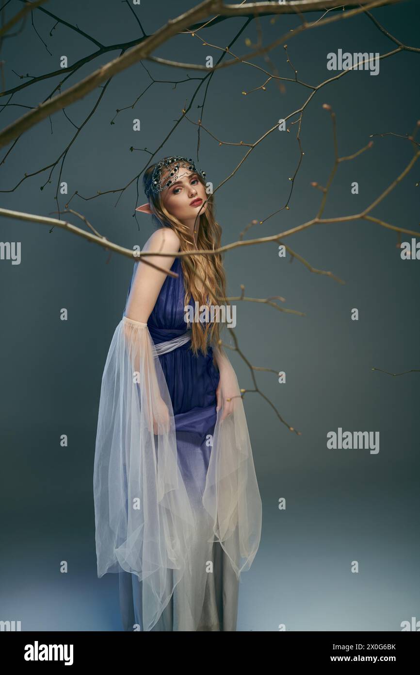 Una giovane donna con un abito blu si erge graziosamente accanto a un albero in un ambiente a tema fato e fantasia. Foto Stock