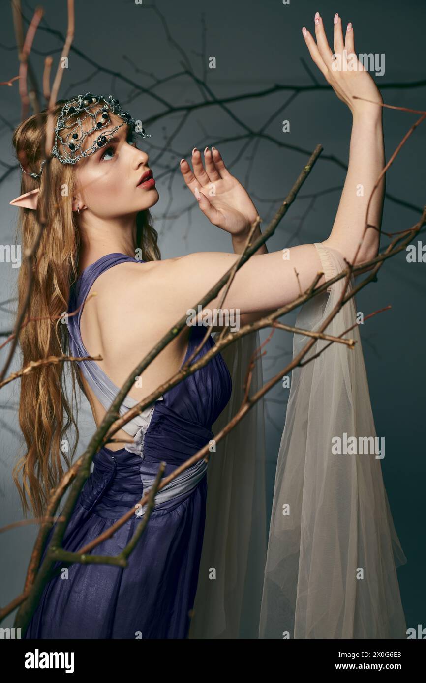 Una giovane donna con un sorprendente abito blu si erge elegantemente di fronte a un maestoso albero in un ambiente da studio, incarnando una principessa elfa. Foto Stock