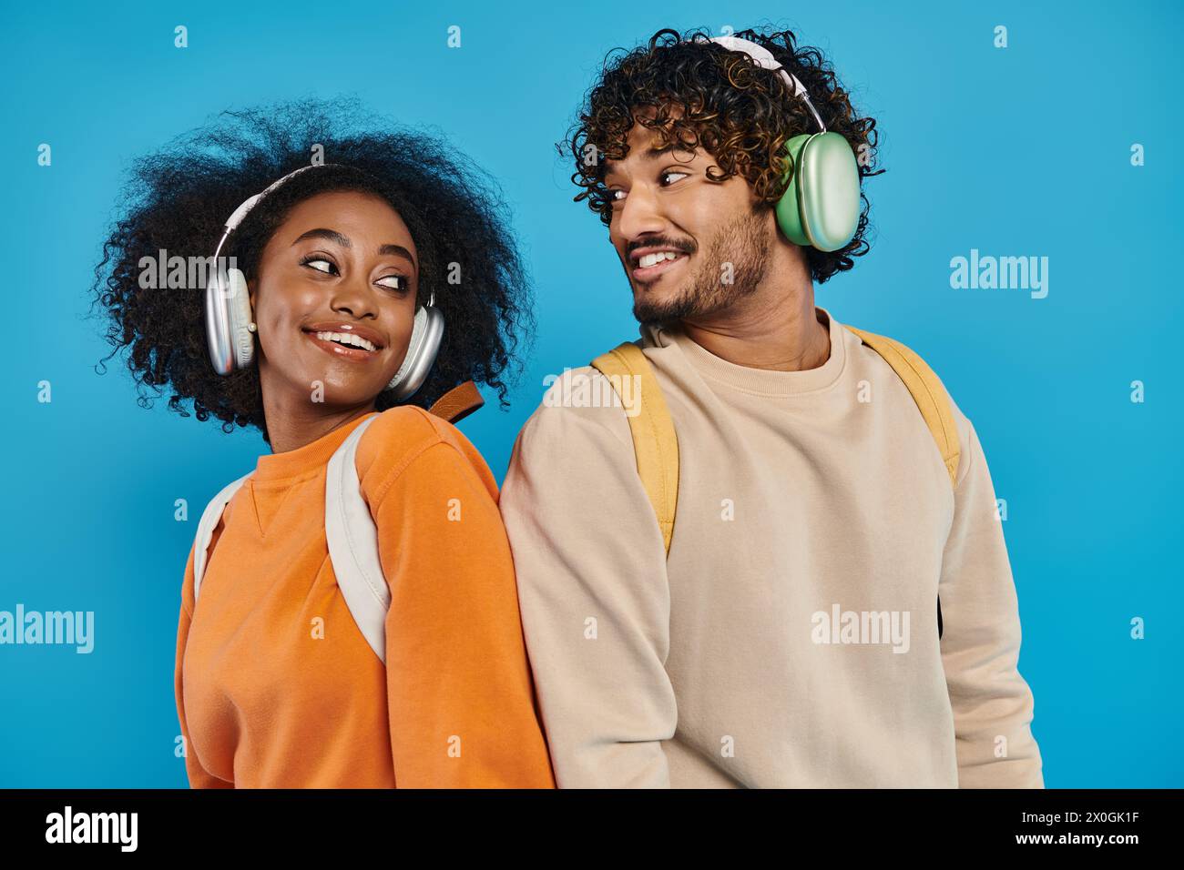 Una coppia interrazziale sta insieme, indossando le cuffie, condividendo la musica su uno sfondo blu in un ambiente da studio. Foto Stock