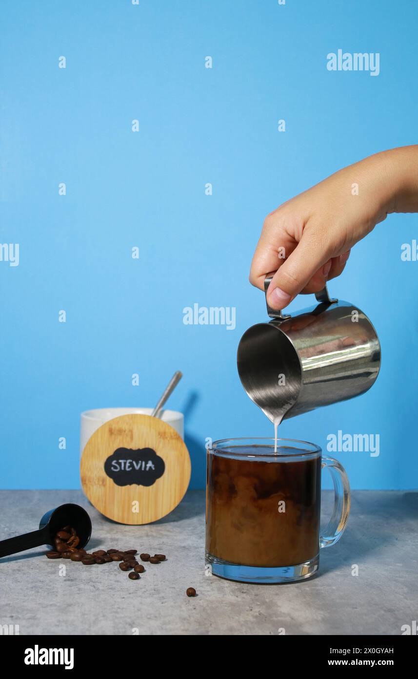 Versare il latte nel caffè nero in un bicchiere, stevia sullo sfondo Foto Stock