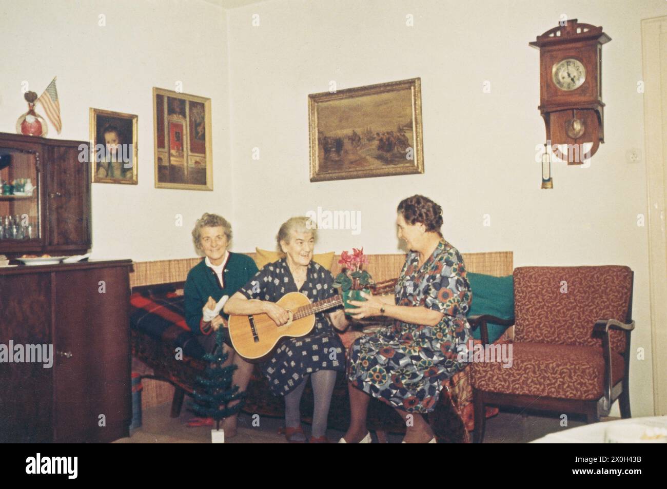 In un soggiorno tre donne in abiti sono seduti su un divano. Uno ha un bendaggio sul suo dito (1° da sinistra), uno suona una chitarra (2° da sinistra) e il terzo tiene una poinsettia nelle sue mani (3° da sinistra). [traduzione automatica] Foto Stock