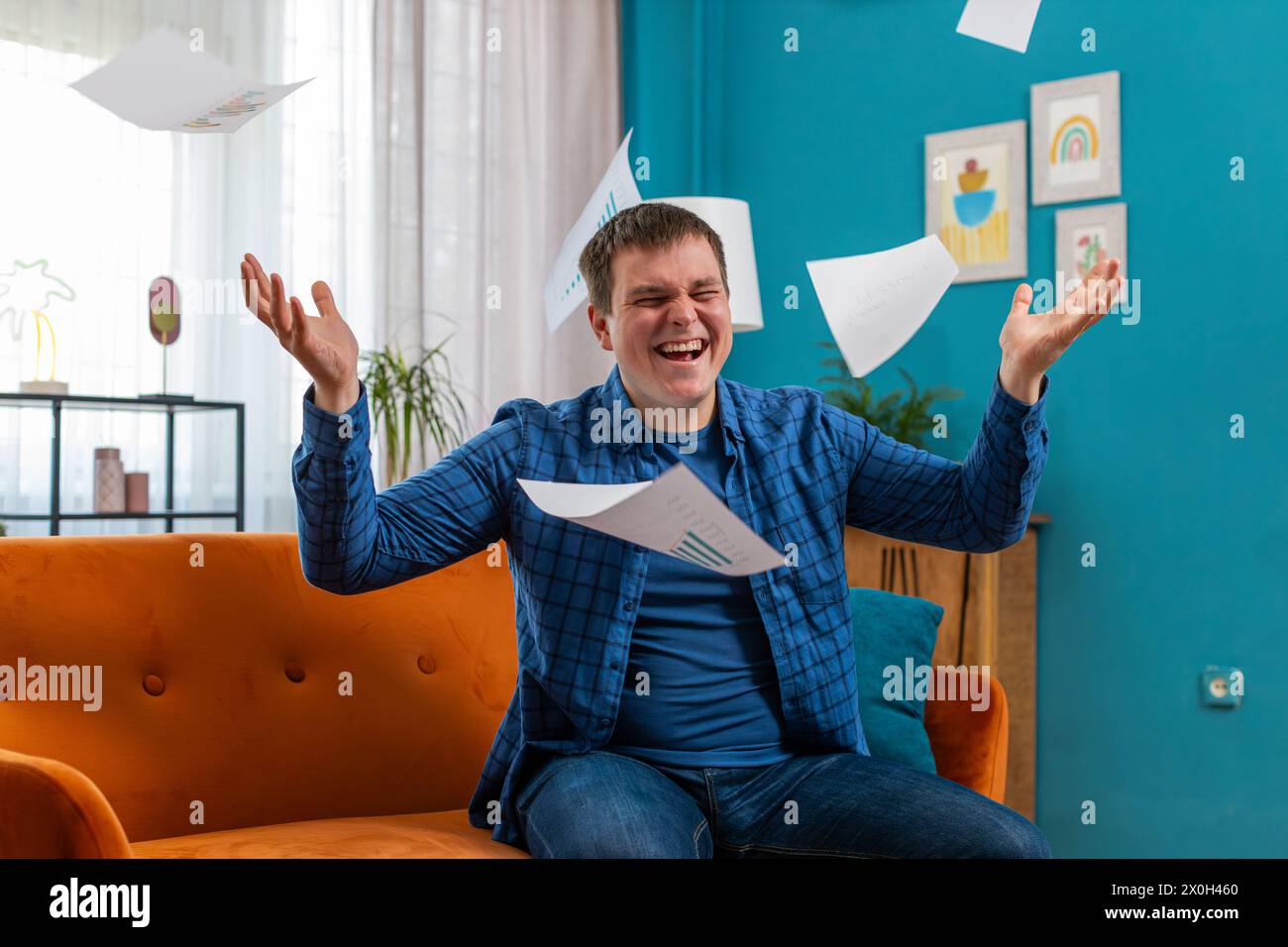 Stressato giovane freelance caucasico che esamina grafici e grafici aziendali e lancia in aria carte non pagate, seduti sul divano nel salotto di casa. Un tizio arrabbiato lavora a distanza sul divano dell'appartamento Foto Stock