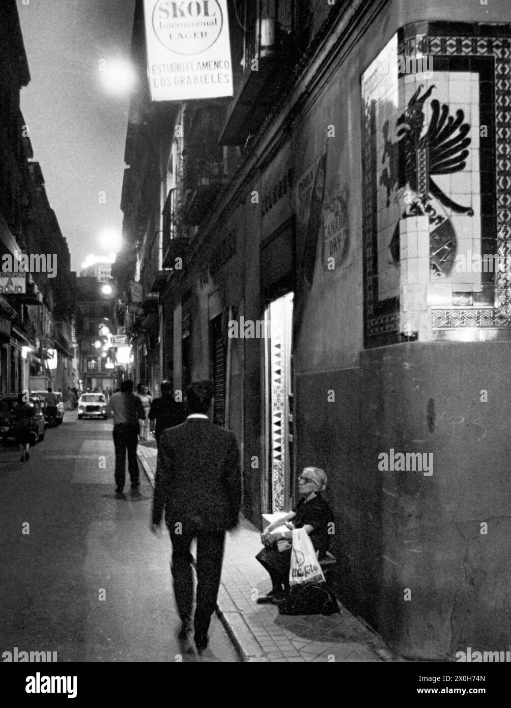 Vista di un vicolo di Madrid di notte. Una donna anziana siede a terra di fronte ad un bar di flamenco. [traduzione automatizzata] Foto Stock