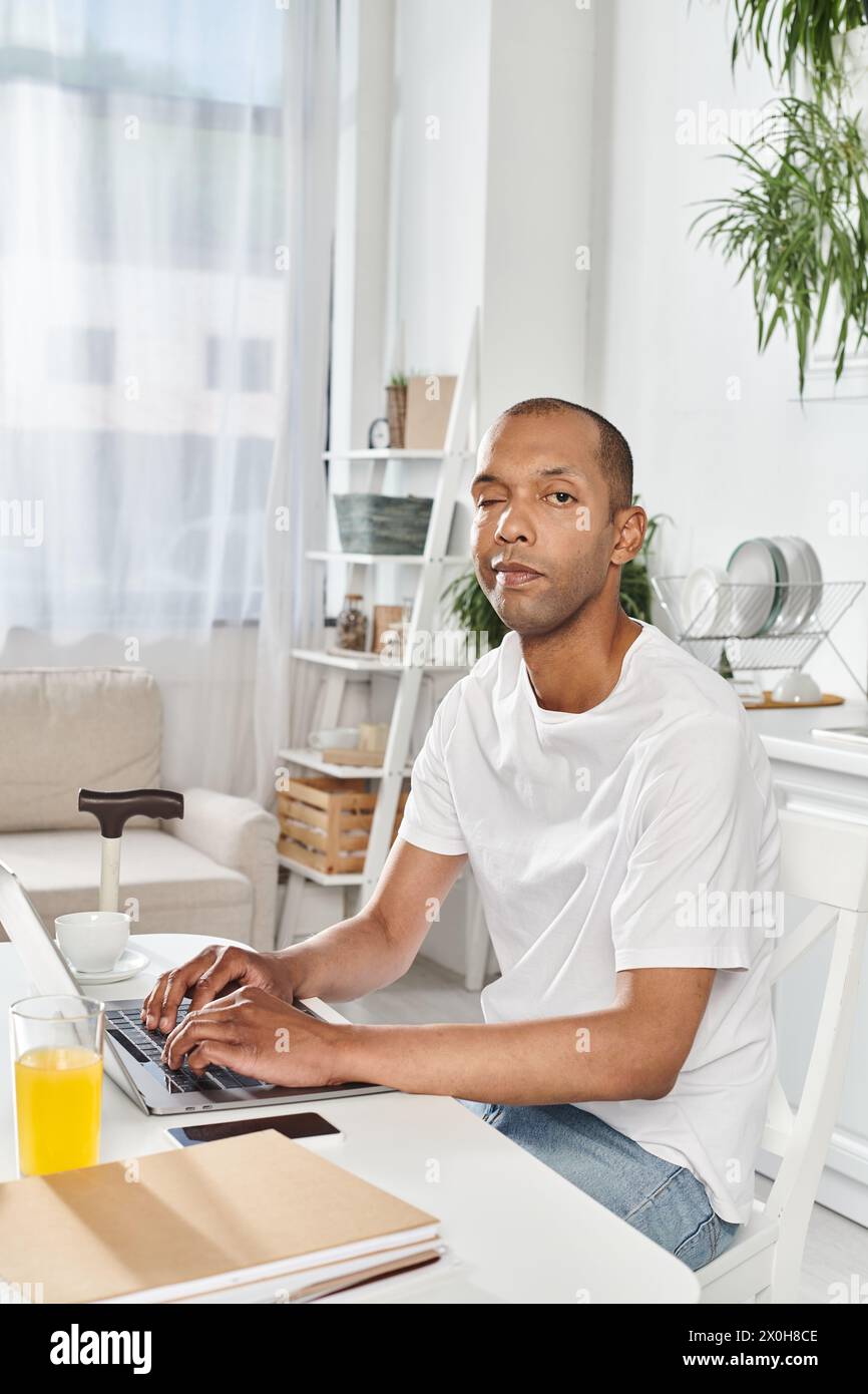 Un uomo di origine afroamericana, che vive con la miastenia gravis, è impegnato nell'uso di un computer portatile a un tavolo. Foto Stock