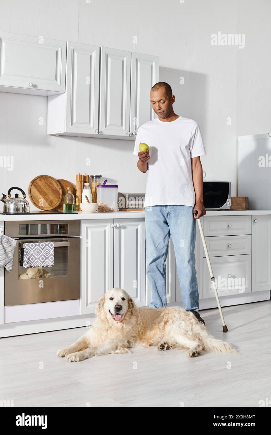 Un afroamericano con miastenia gravis in piedi in una cucina con un bastone e il suo leale cane Labrador. Foto Stock
