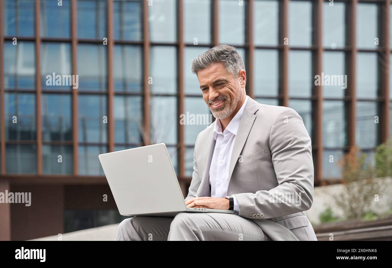 Uomo d'affari di mezza età felice e impegnato che lavora su un notebook fuori dall'ufficio. Foto Stock