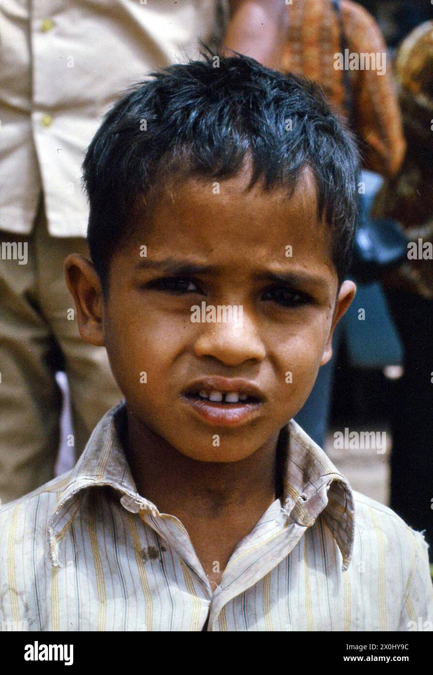 Ritratto di un ragazzino in India. Ha capelli neri corti e indossa una camicia colorata a righe. Stringe, accecato dal sole, gli occhi un po' chiusi dietro di lui un uomo e una donna. Indossa un sari. [traduzione automatica] Foto Stock