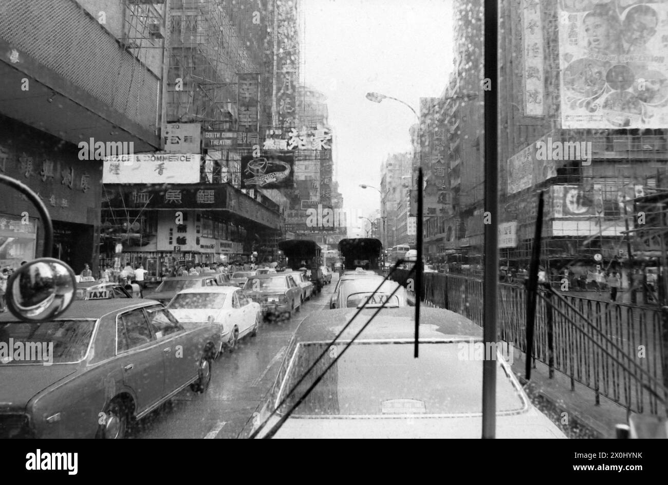 Vista lungo una strada a Hong Kong sotto la pioggia. La strada è molto trafficata e ci sono grattacieli a sinistra e a destra. I grattacieli sono coperti da grandi poster pubblicitari. La foto è stata scattata attraverso il parabrezza di un autobus. È possibile vedere uno specchietto retrovisore e i tergicristalli. [traduzione automatica] Foto Stock