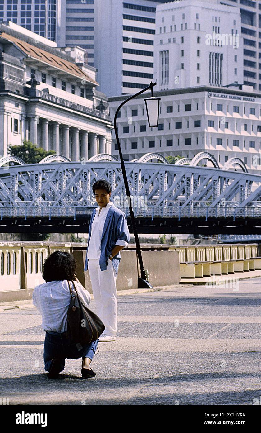 Un uomo posa per una foto su una passeggiata di fronte ad un paesaggio impressionante a Singapore. Indossa pantaloni bianchi, una camicia bianca, una camicia blu e mette le mani in tasca. Una donna fotografa la scena, inginocchiandosi a terra, indossando jeans, una blusa bianca e una borsa sulla spalla. Sullo sfondo si trova un ponte bianco in ferro battuto, una lampada a strisce, sulla sinistra un edificio neoclassico e altri grattacieli e grattacieli moderni. [traduzione automatica] Foto Stock