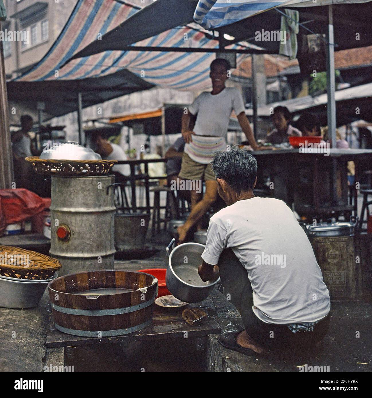 Un ristorante di strada (Hawker) a Hong Kong. In primo piano un uomo è seduto in posizione di accovacciamento e lavando una pentola. Un altro uomo lo guarda ridere. Dietro di lui ci sono tavoli con ombrelloni di uno snack bar sulla strada. Alcune persone stanno mangiando loro. [traduzione automatica] Foto Stock