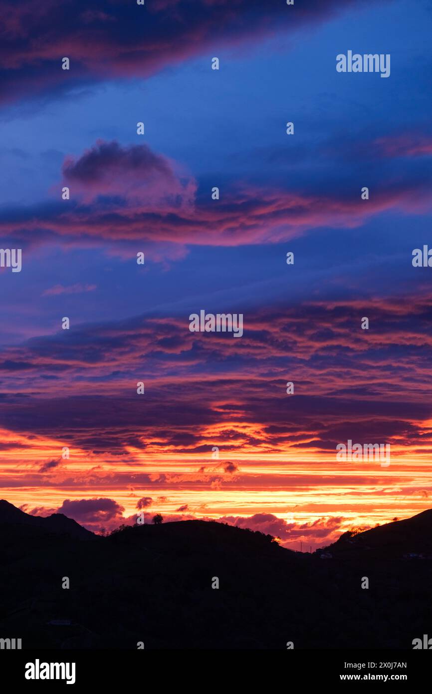 Il sole tramonta dietro uno sfondo di nuvole e montagne torreggianti, che proiettano un caldo bagliore sul paesaggio. Foto Stock