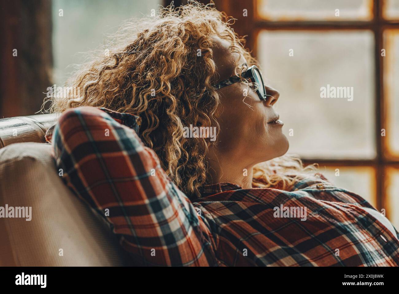 Donna adulta di mezza età che si allunga e si rilassa a casa seduta sulla sedia guardando fuori dalle finestre sullo sfondo - concetto di stile di vita femminile single felice - primo piano di giovane maturo con gli occhiali da vista Foto Stock
