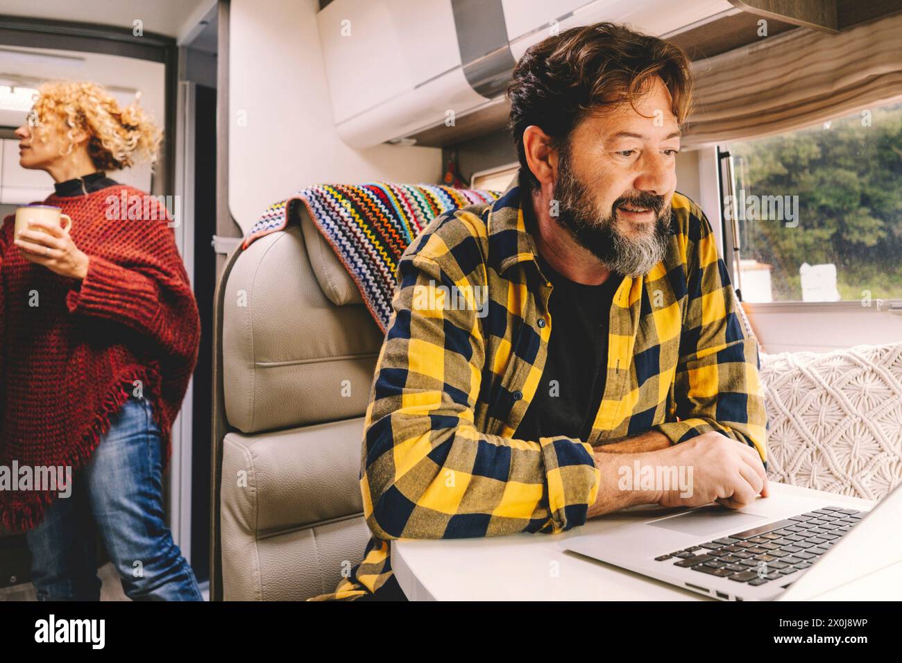 Una coppia di adulti viaggia e si diverte a campeggiare in auto per vacanze o per vivere la vita d'avanguardia - un uomo felice lavora su un computer portatile sul tavolo e una donna in cerca di oggetti all'interno del veicolo - natura all'aperto Foto Stock