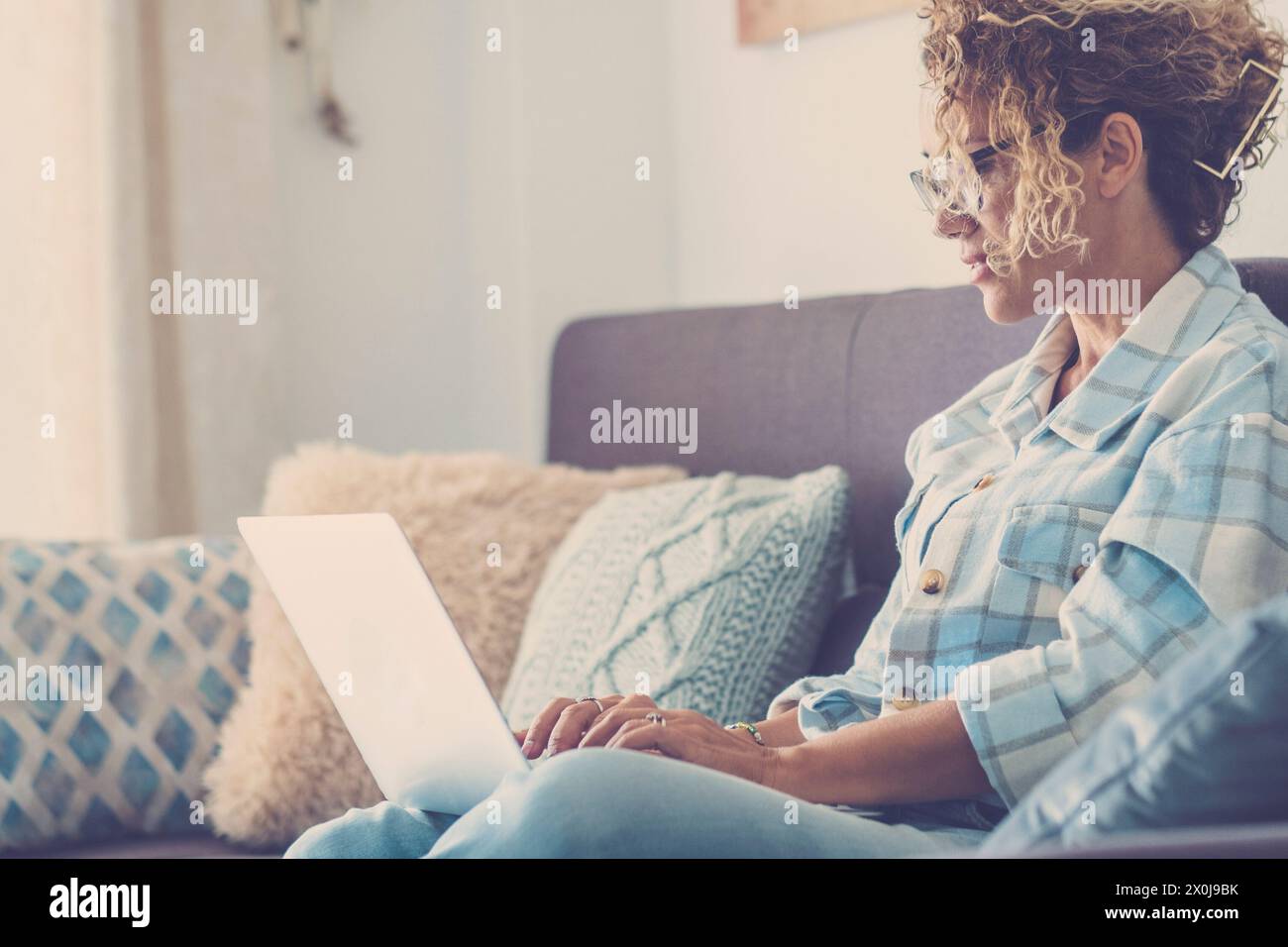 Una moderna donna adulta hipster utilizza il computer portatile a casa, seduto sul divano, e si rilassa nelle attività al chiuso. Una donna al lavoro online. Signora a casa seduta sul divano Foto Stock