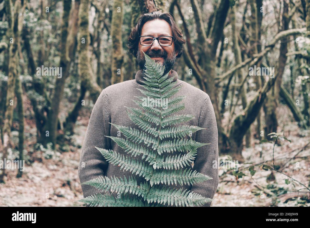 Un uomo maturo sorride e guarda la macchina fotografica con grandi foglie tropicali verdi e foresta di alberi sullo sfondo. Persone e natura nelle attività ricreative all'aperto. Stile di vita bio sano alternativo. Ambiente Foto Stock