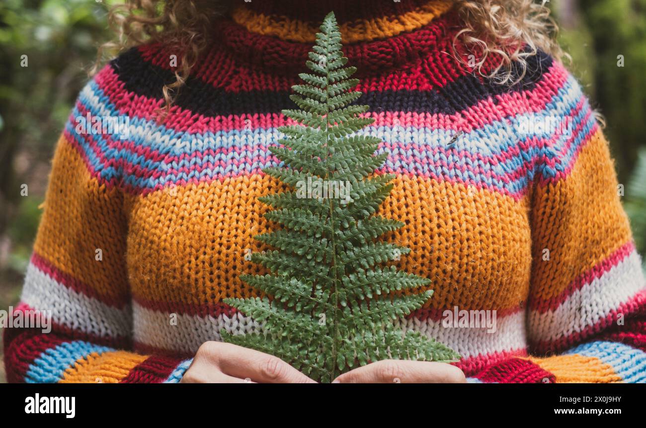 Ritaglia il ritratto di una donna irriconoscibile in natura, indossando un maglione colorato e tenendo una piccola foglia tropicale contro il cuore e il corpo. Persone che amano la natura all'aperto. Viaggiare stile di vita sano Foto Stock