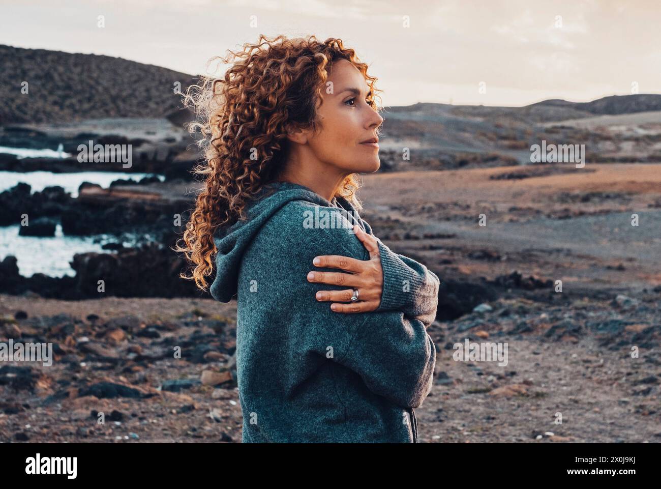 Una donna sola in campagna all'aperto si abbraccia e sorride con espressione serena guardando lontano. Mulini a vento e montagne sullo sfondo. Destinazione di viaggio e viaggiatori con stile di vita adulto Foto Stock