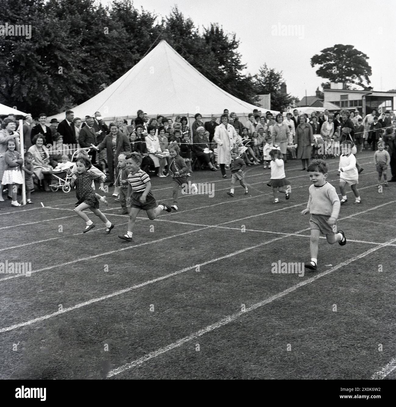 1964, bambini storici che partecipano a gare di corsa all'aperto su un circuito erboso in occasione di una giornata di sport e attività della comunità, Inghilterra, Regno Unito. Foto Stock