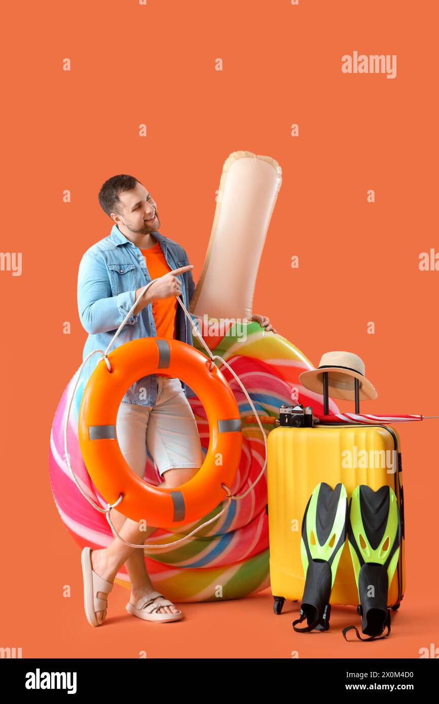 Giovane uomo con materasso gonfiabile a forma di caramelle, valigia e accessori da spiaggia che puntano a qualcosa su sfondo arancione Foto Stock