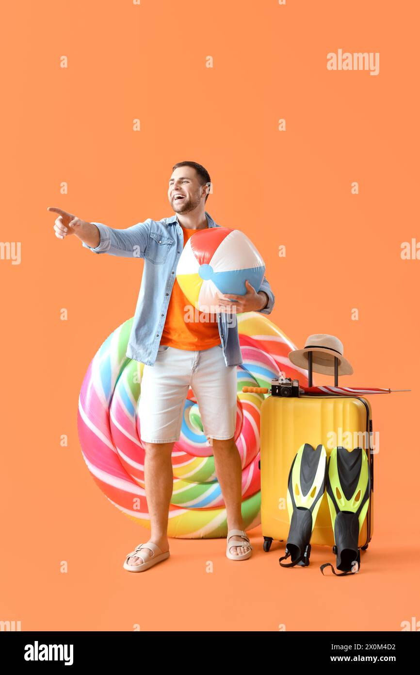 Giovane uomo con materasso gonfiabile a forma di caramelle, valigia e accessori da spiaggia che puntano a qualcosa su sfondo arancione Foto Stock