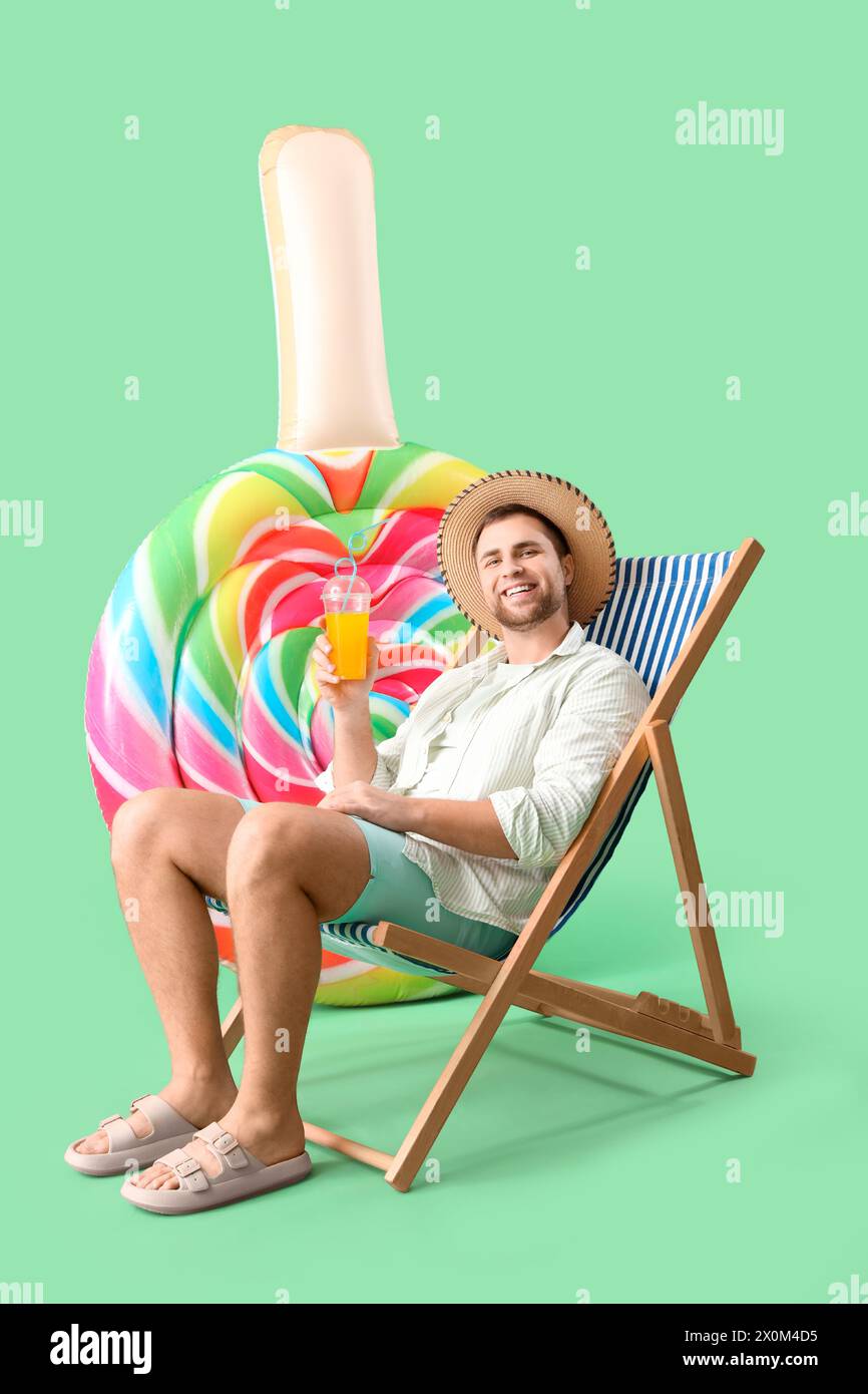 Giovane uomo con materasso gonfiabile a forma di caramelle seduto su sdraio su sfondo verde Foto Stock