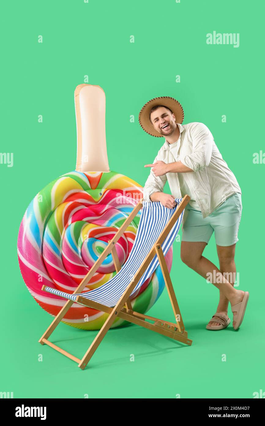 Giovane uomo con materasso gonfiabile a forma di caramelle seduto su sdraio su sfondo verde Foto Stock