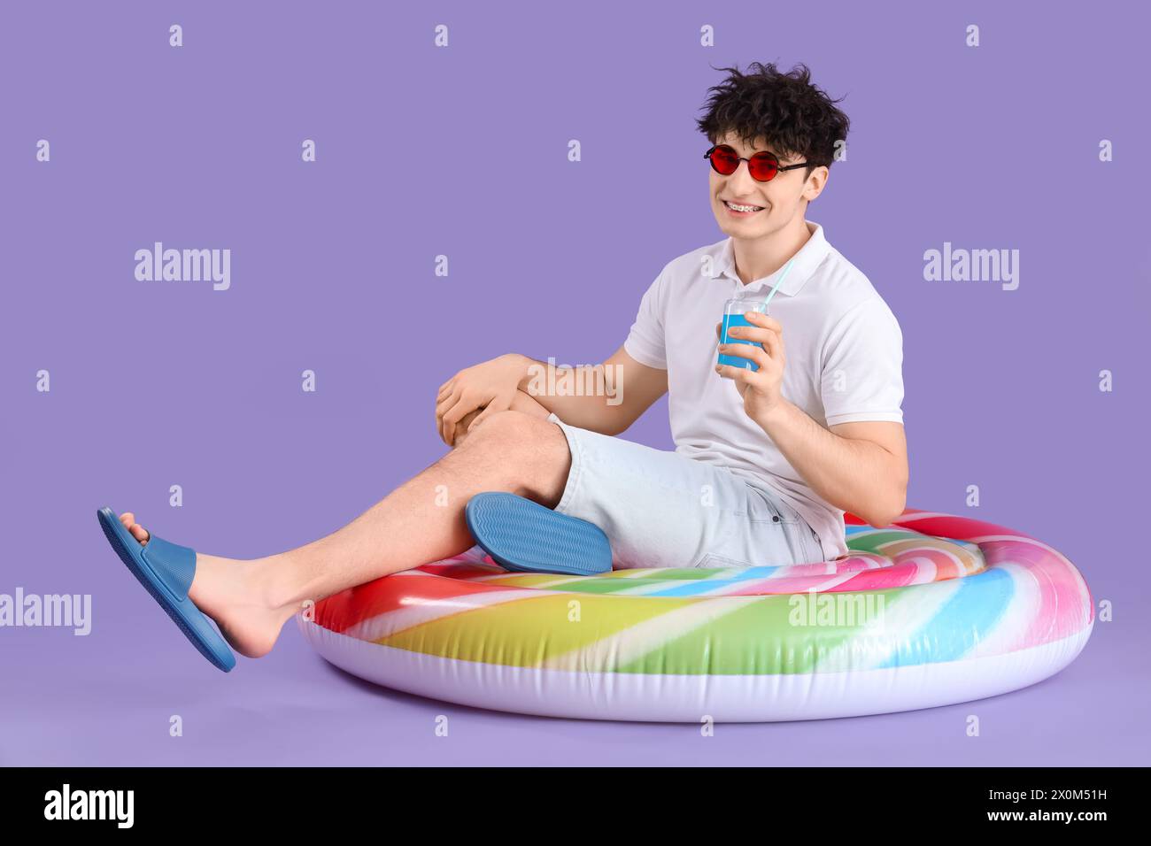 Bel giovane uomo felice con materasso gonfiabile e bicchiere di cocktail su sfondo viola Foto Stock