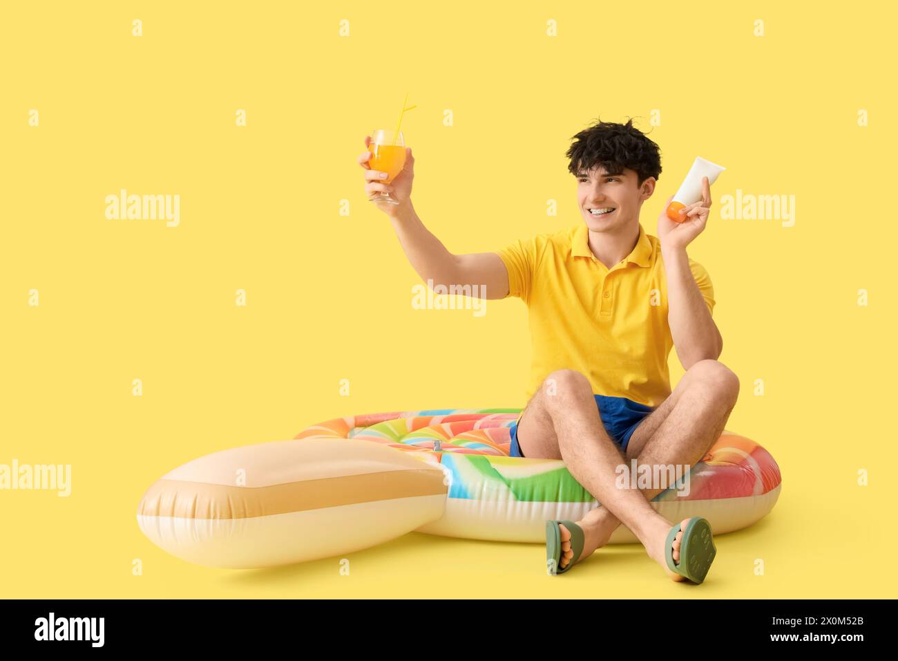 Bel giovane uomo felice con materasso gonfiabile, crema solare e bicchiere di succo d'arancia su sfondo giallo Foto Stock