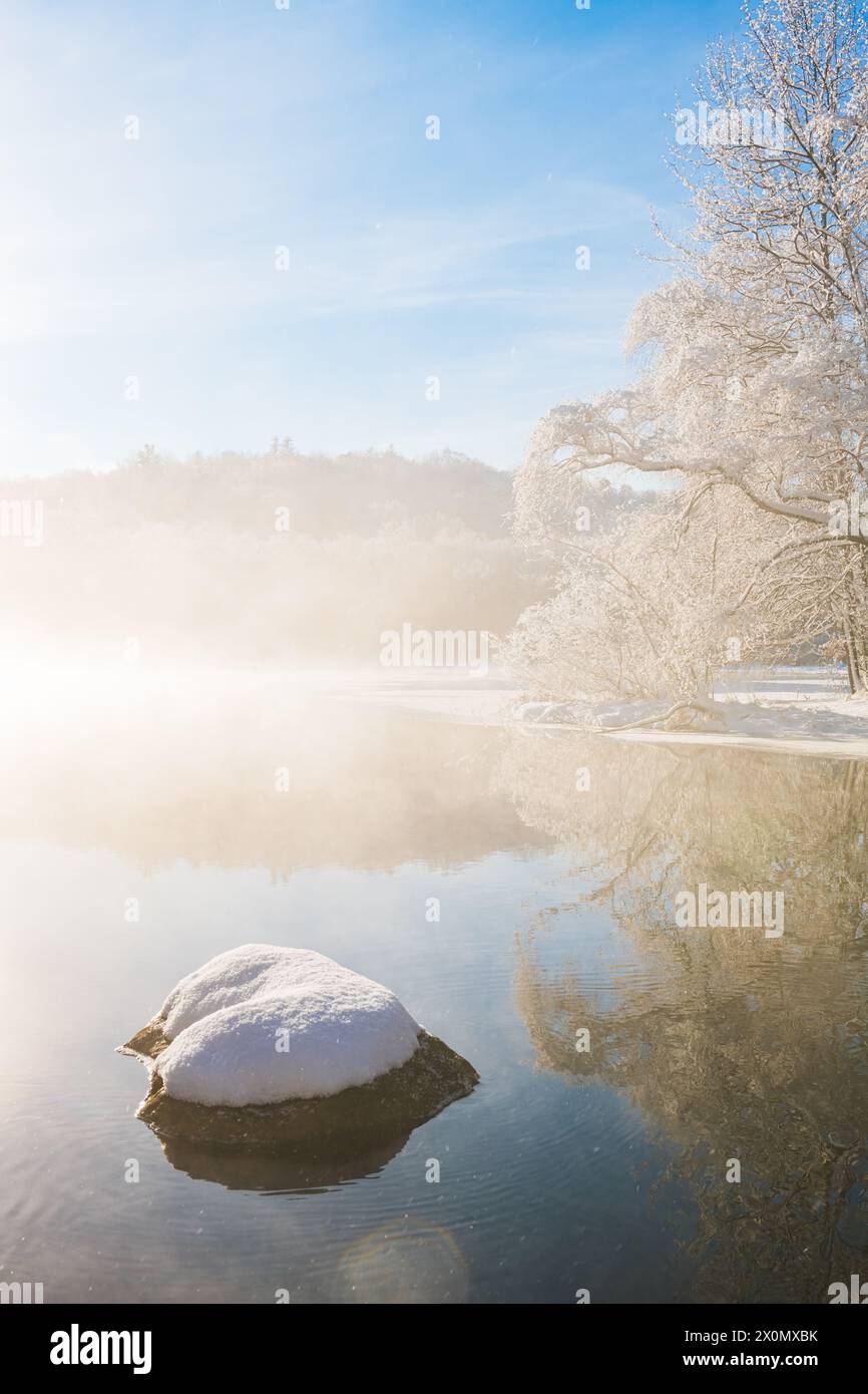 A Mölndal, in Svezia, si apre una tranquilla mattina d'inverno, mentre il sole nascente penetra attraverso la nebbia sopra il fiume calmo. La neve ricopre le rocce e gli alberi Foto Stock