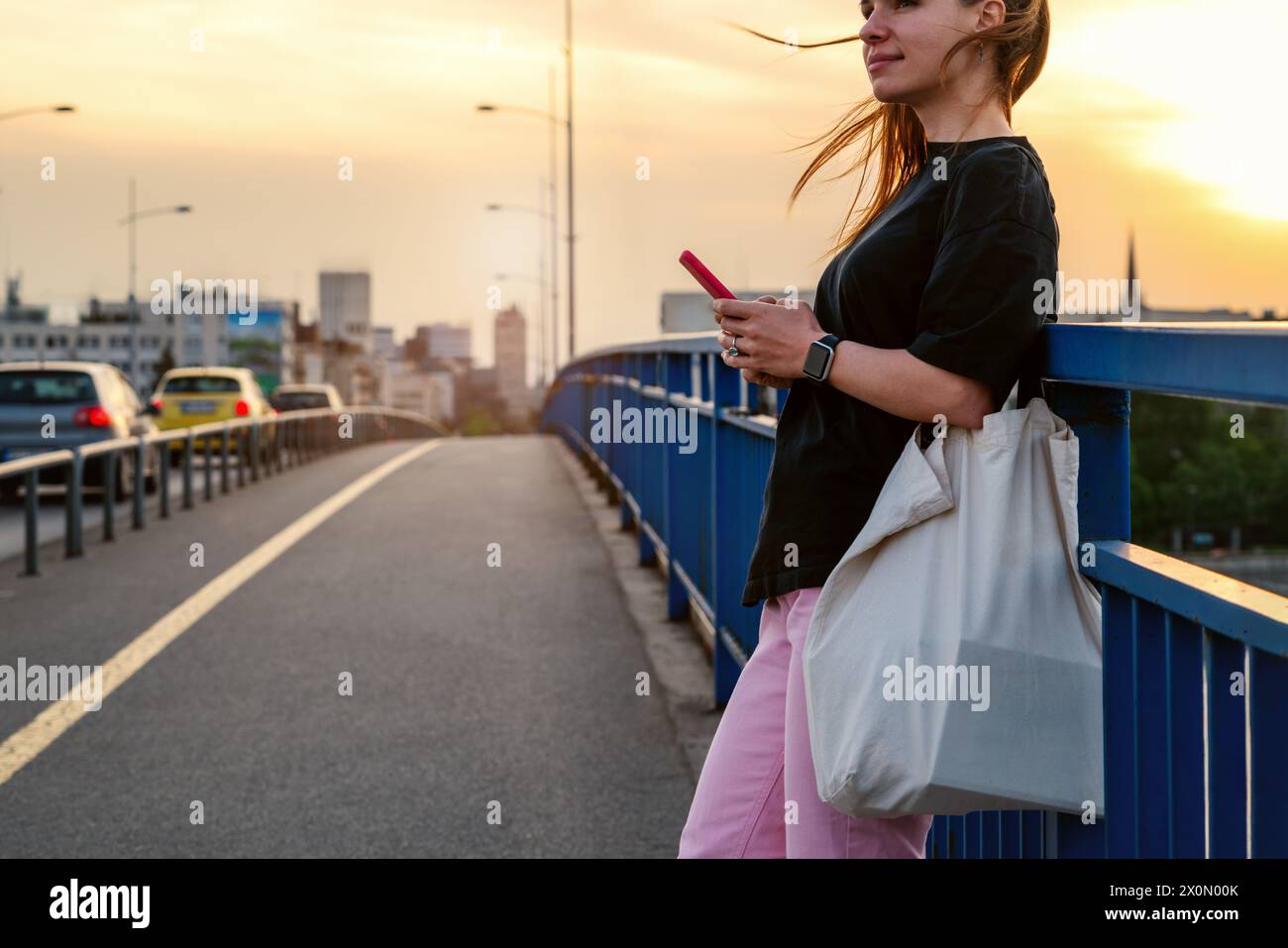 Stile di vita cittadino. Donna urbana che indossa un outfit casual sta sul ponte e utilizza il suo smartphone. Foto Stock