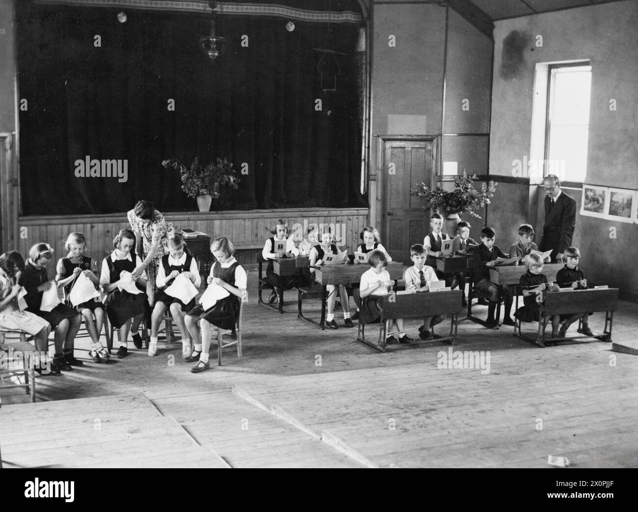 LONDRA EVACUATI NEL CARMARTHENSHIRE, GALLES, 1940 - i bambini della Woodmansterne Road School di Streatham, Londra frequentano le lezioni tenute nella sala del villaggio di Farmers, Carmarthenshire, Galles nel 1940. Le ragazze più grandi usano un'area della sala per la loro lezione di cucito, mentre altri bambini più piccoli leggono e lavorano a maglia presso i banchi di un'altra area della sala. Entrambi i gruppi sono supervisionati da un insegnante separato Foto Stock