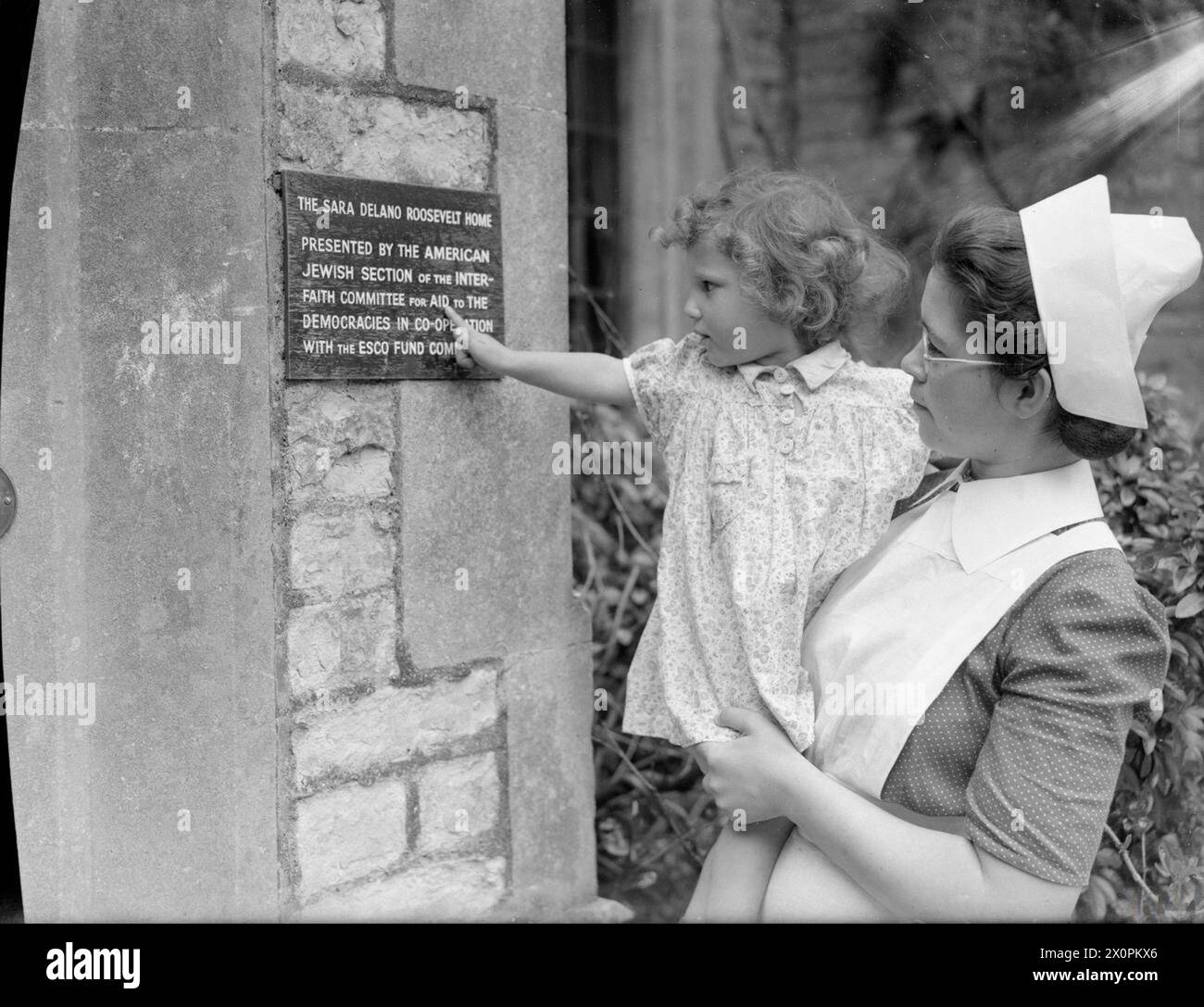 AMERICAN AID TO BRITAIN: LIFE AT SYSTON COURT NURSERY, THE SARA DELANO ROOSEVELT HOME, GLOUCESTERSHIRE, INGHILTERRA, 1942 - Un'infermiera tiene un bambino in modo che possa leggere la targa del nome della nursery in cui risiede. La targa recita: "The Sara Delano Roosevelt Home. Presentato dalla sezione ebraica americana del Comitato interreligioso per gli aiuti alle democrazie in cooperazione con il Comitato del Fondo esco" Foto Stock