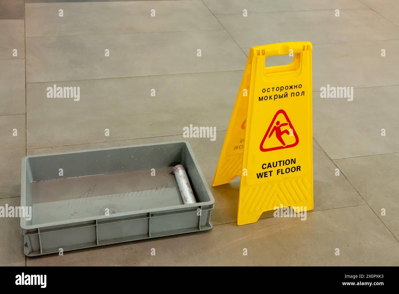 Accanto a un vassoio d'acqua in plastica grigia è presente un cartello giallo per il pavimento bagnato Un panno sporco su un pavimento piastrellato, per garantire che le misure di sicurezza siano applicate Foto Stock