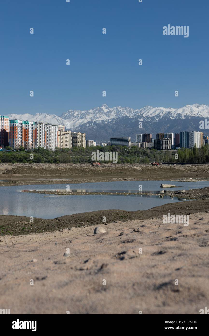 Serbatoio di stoccaggio Lago Sayran, Almaty, Kazakistan. Spiaggia di sabbia di Empty City con laghetto drenato. Edifici residenziali e alta cima innevata Foto Stock