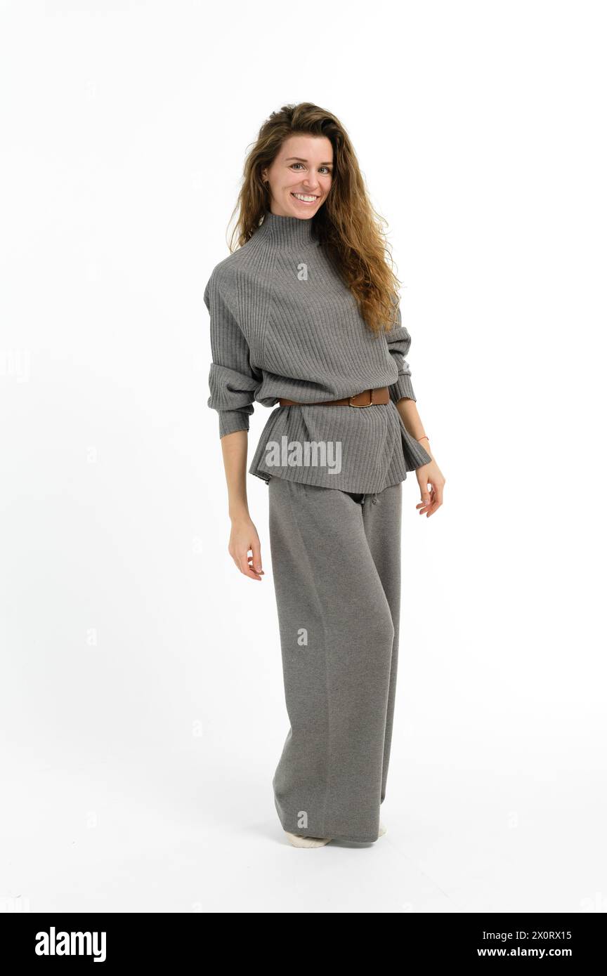 Una donna con un sorriso radioso, indossa un elegante pullover grigio e pantaloni a gamba larga. I suoi capelli cadono giù per il collo, le maniche arrotolate, trasudando confidenza Foto Stock