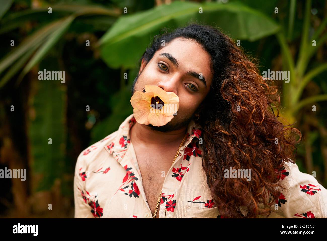 Uomo dell'Asia meridionale con fiore in bocca posa, mostrando trucco espressivo, audace femminilità. Indossando una camicia floreale in un fogliame verde, il genere è rappresentato Foto Stock