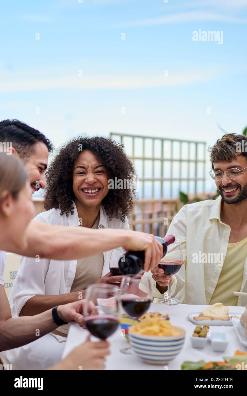 Giovane uomo caucasico sorridente che serve vino rosso agli ospiti al tavolo per festeggiare gli amici felici. Foto Stock