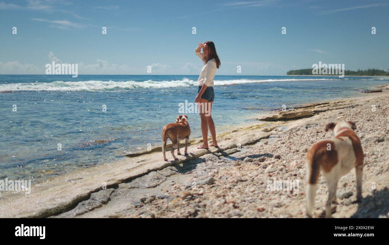 Una donna è in piedi su una spiaggia sabbiosa accanto a due cani. La donna guarda l'oceano mentre i cani giocano nella sabbia. La scena cattura un momento tranquillo di compagnia tra umani e animali. Foto Stock