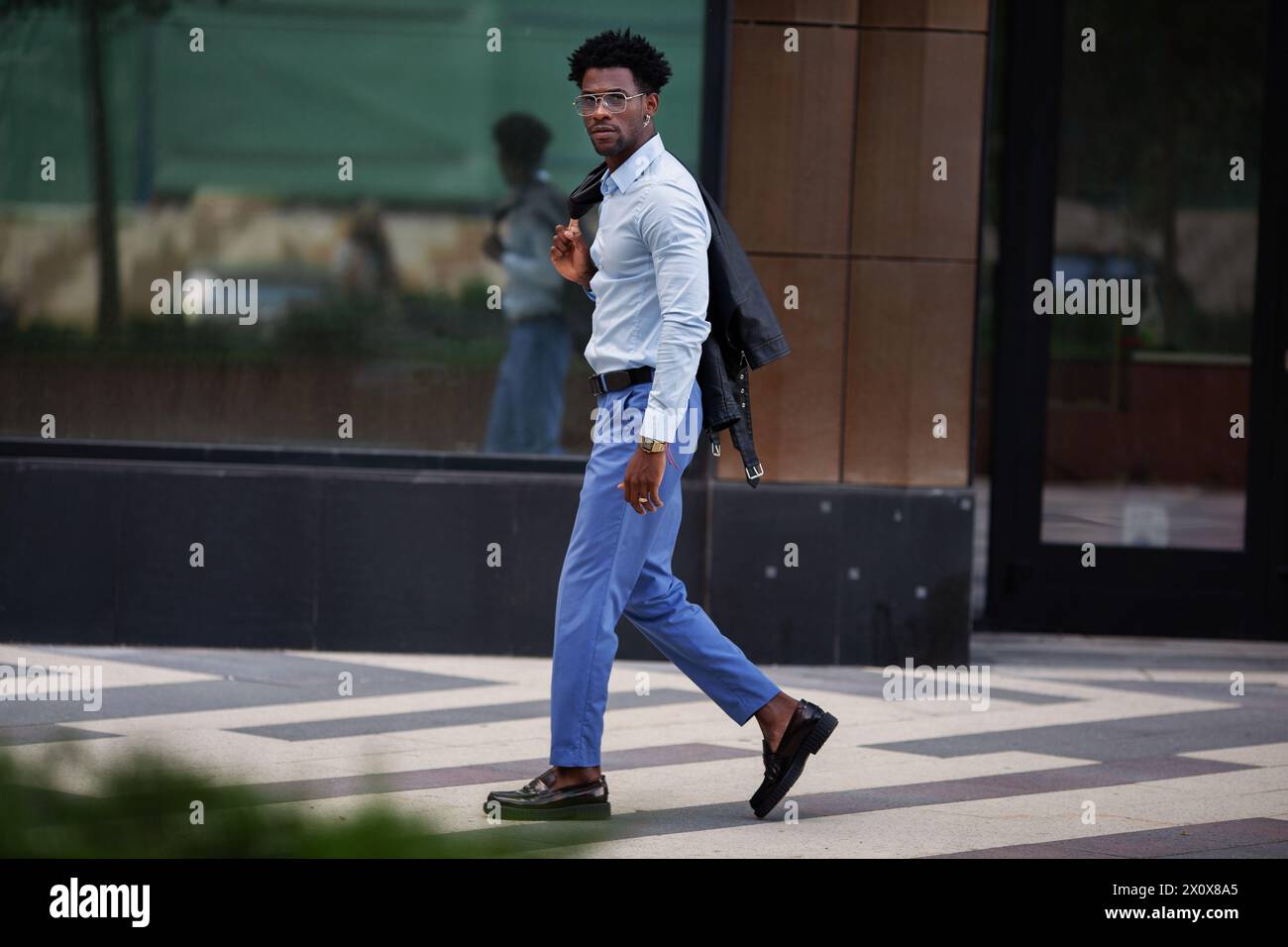 Elegante uomo afroamericano alla moda che cammina all'aperto in città, indossando pantaloni blu e una camicia con mocassini, trasuda raffinatezza urbana. Foto Stock