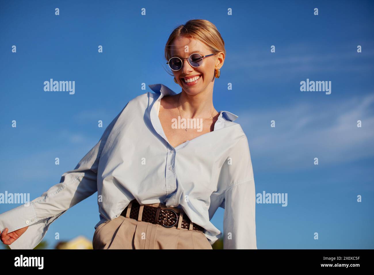Donna contemporanea felice con abiti eleganti, camicia oversize e occhiali da sole, che sorride gioiosamente sul cielo blu. Atmosfera estiva alla moda, felice ed elegante. Foto Stock
