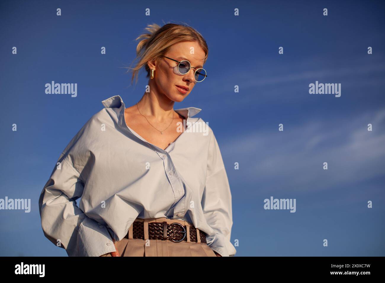 Donna moderna con una camicia oversize e occhiali da sole si staglia all'aperto contro il cielo. Chic, contemporaneo ed elegante, cattura la libertà estiva. Foto Stock