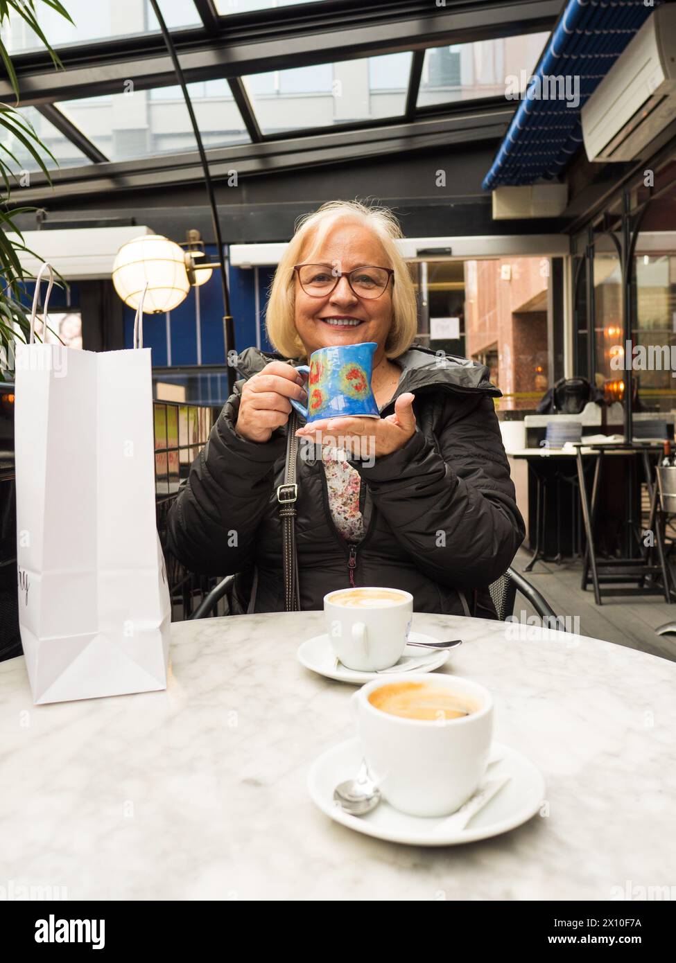 Una donna anziana bionda sorridente mostra un regalo fatto a mano per la festa della mamma in un bar Foto Stock