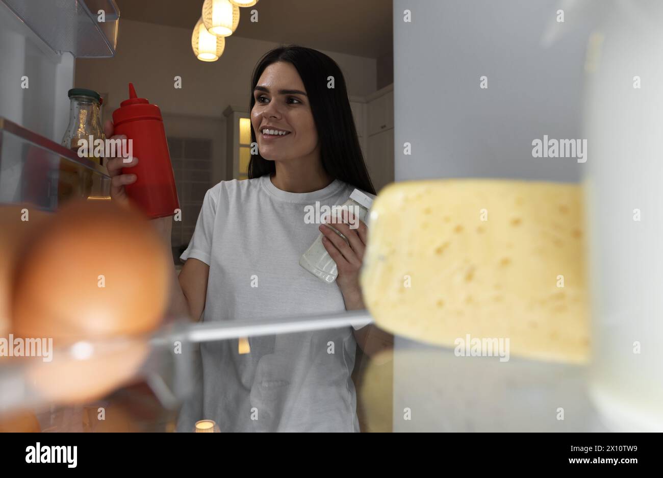 Una giovane donna che porta ketchup e maionese fuori dal frigorifero in cucina di notte, vista dall'interno Foto Stock
