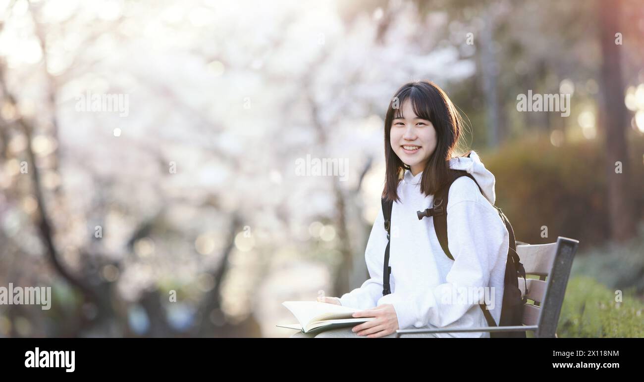 Concetto di uno studente universitario seduto su una panchina in un parco scolastico, leggendo un libro e studiando, e uno studente delle scuole superiori sorridente mentre porta con sé una borsa Foto Stock