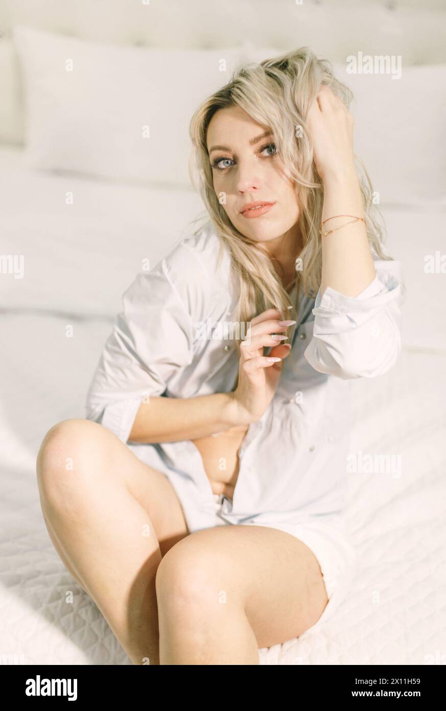 Una donna è seduta sopra un letto bianco, mostrando un'estetica semplice e pulita in una camera da letto. Foto Stock