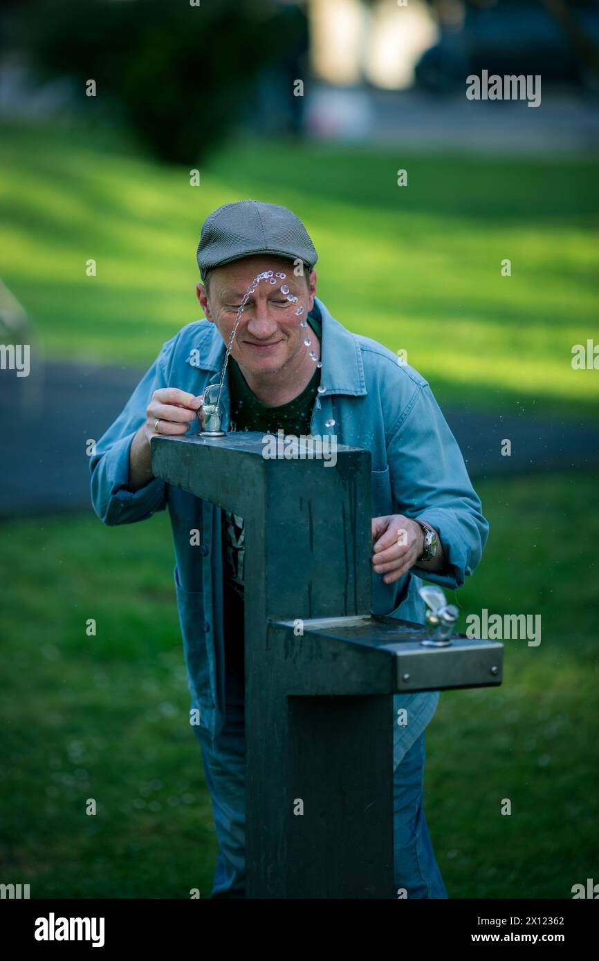 Un uomo disseta dalla fontana di un parco, rinfrescandosi in una giornata calda mentre si ferma per godersi i semplici piaceri della vita. Foto Stock
