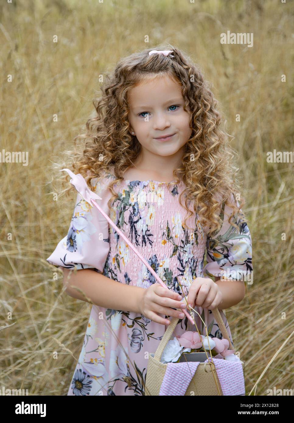 Bella ragazza dai capelli ricci vestita con un abito rosa che regge l'estate a forma di stella e una borsa in un campo coltivato con erba marrone pastello Foto Stock