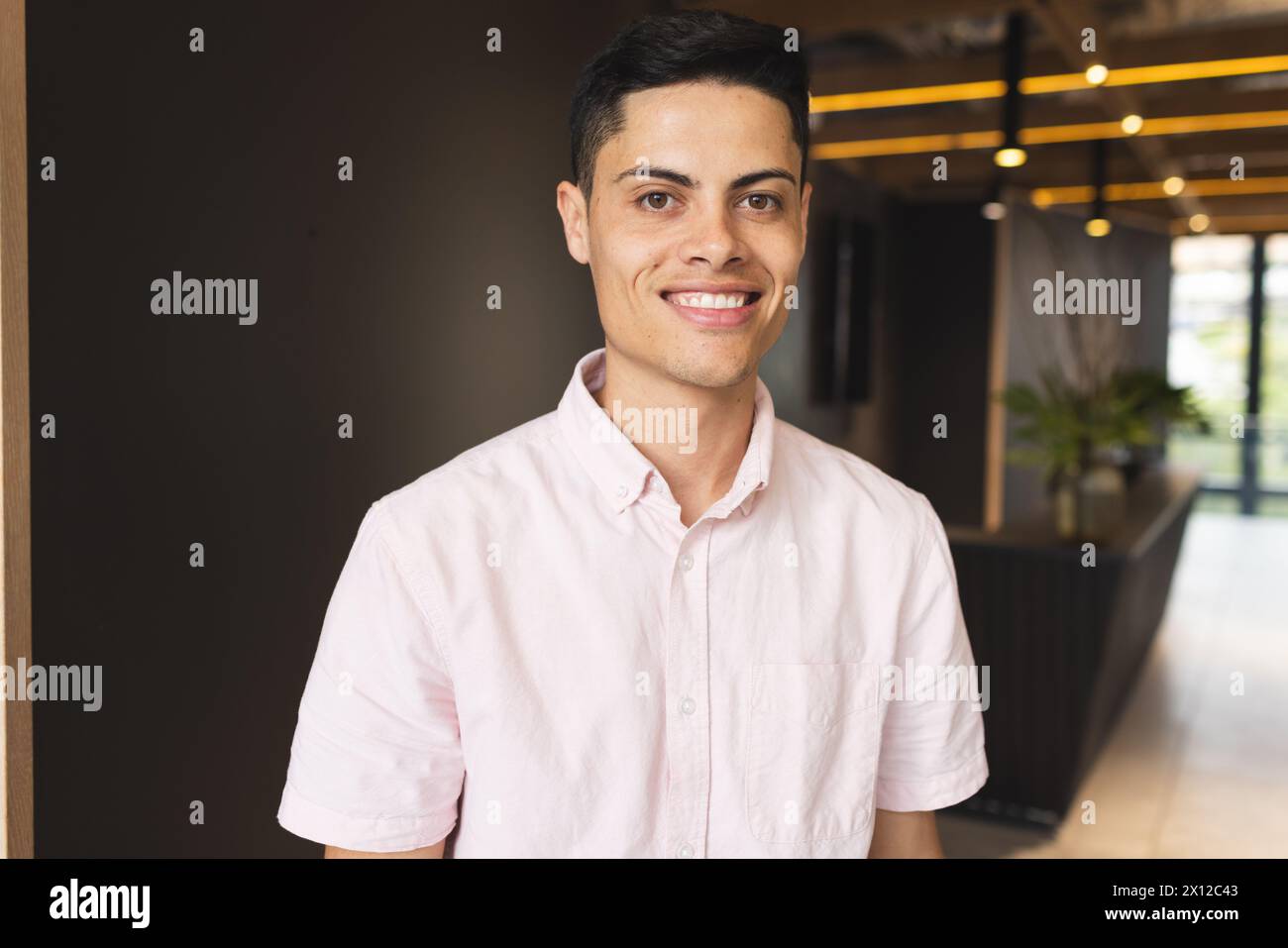 Un giovane uomo birazziale che indossa una camicia rosa sorride in un moderno ufficio aziendale. Ha capelli scuri corti, pelle marrone chiaro e occhi marroni, inalterati. Foto Stock