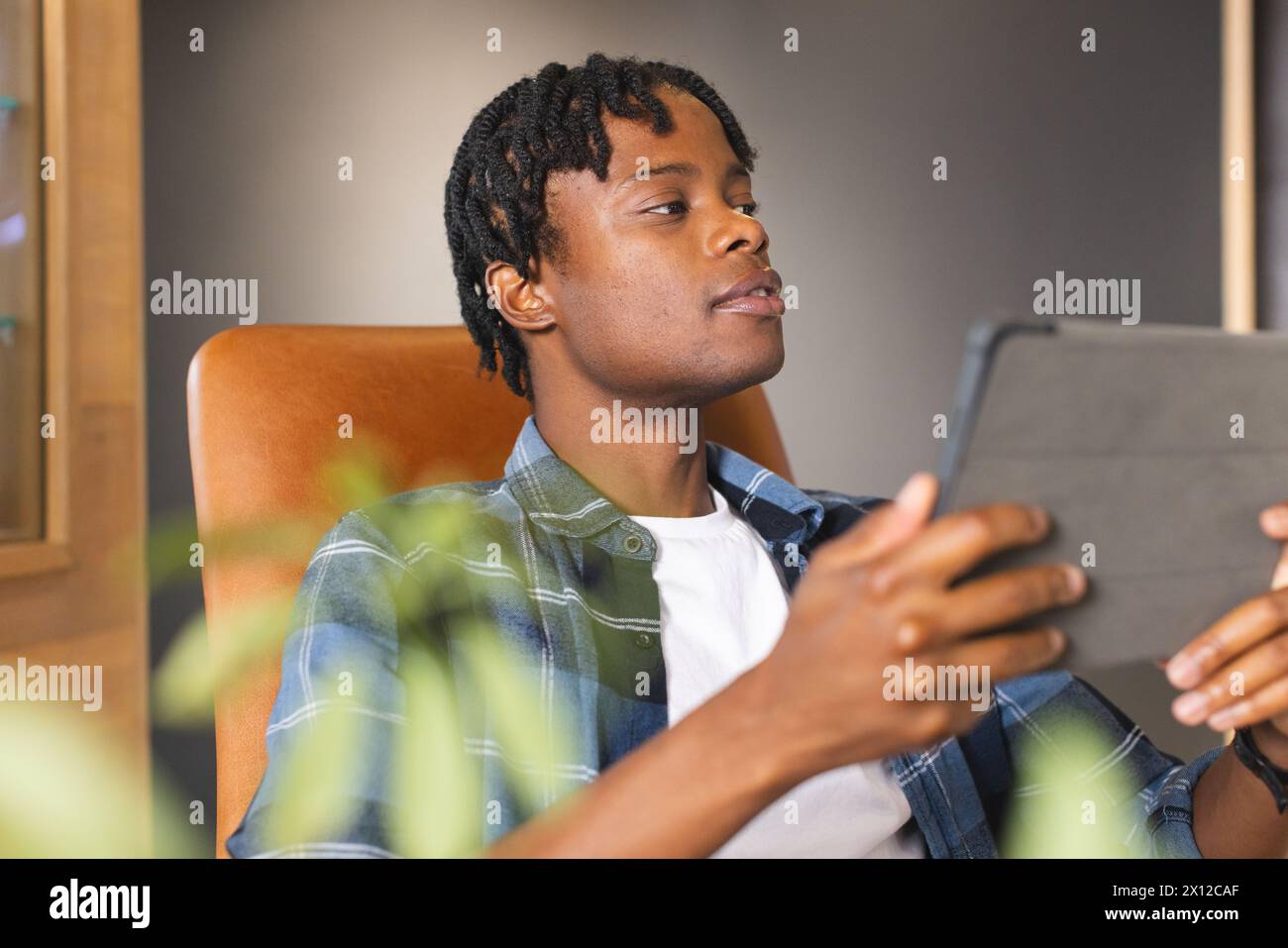 Uomo afroamericano che tiene un tablet, guarda lo schermo in un moderno ufficio aziendale. Ha i capelli corti intrecciati, indossa una camicia a quadri, rilassato su una sedia Foto Stock